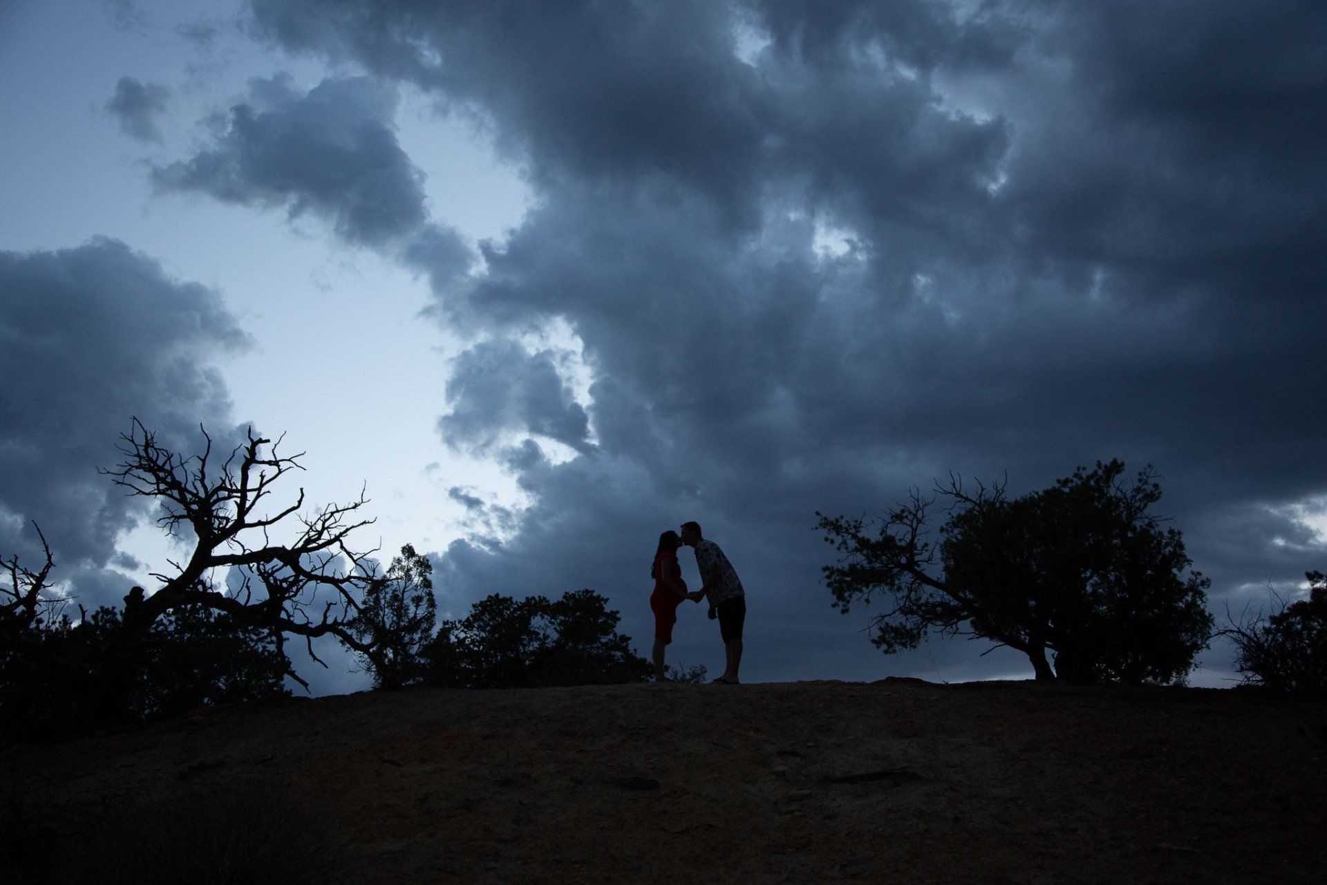 A couple kissing on top of a hill under a cloudy sky.