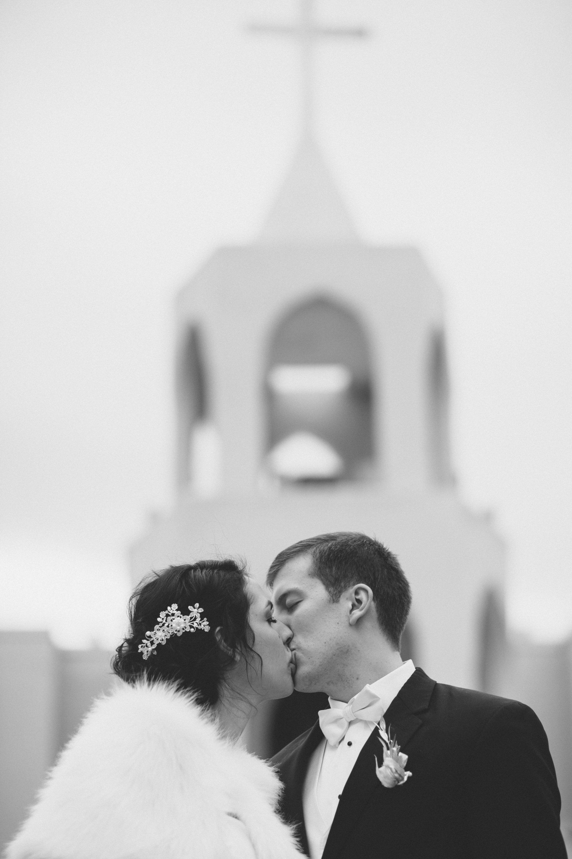 A bride and groom are kissing in front of a church tower.