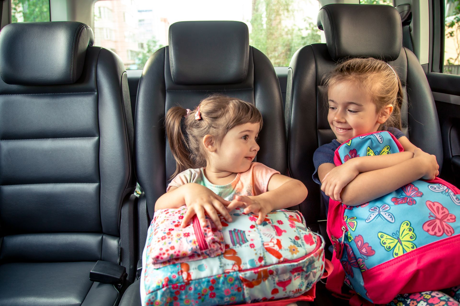 Two little girls are sitting in the back seat of a car with their backpacks.