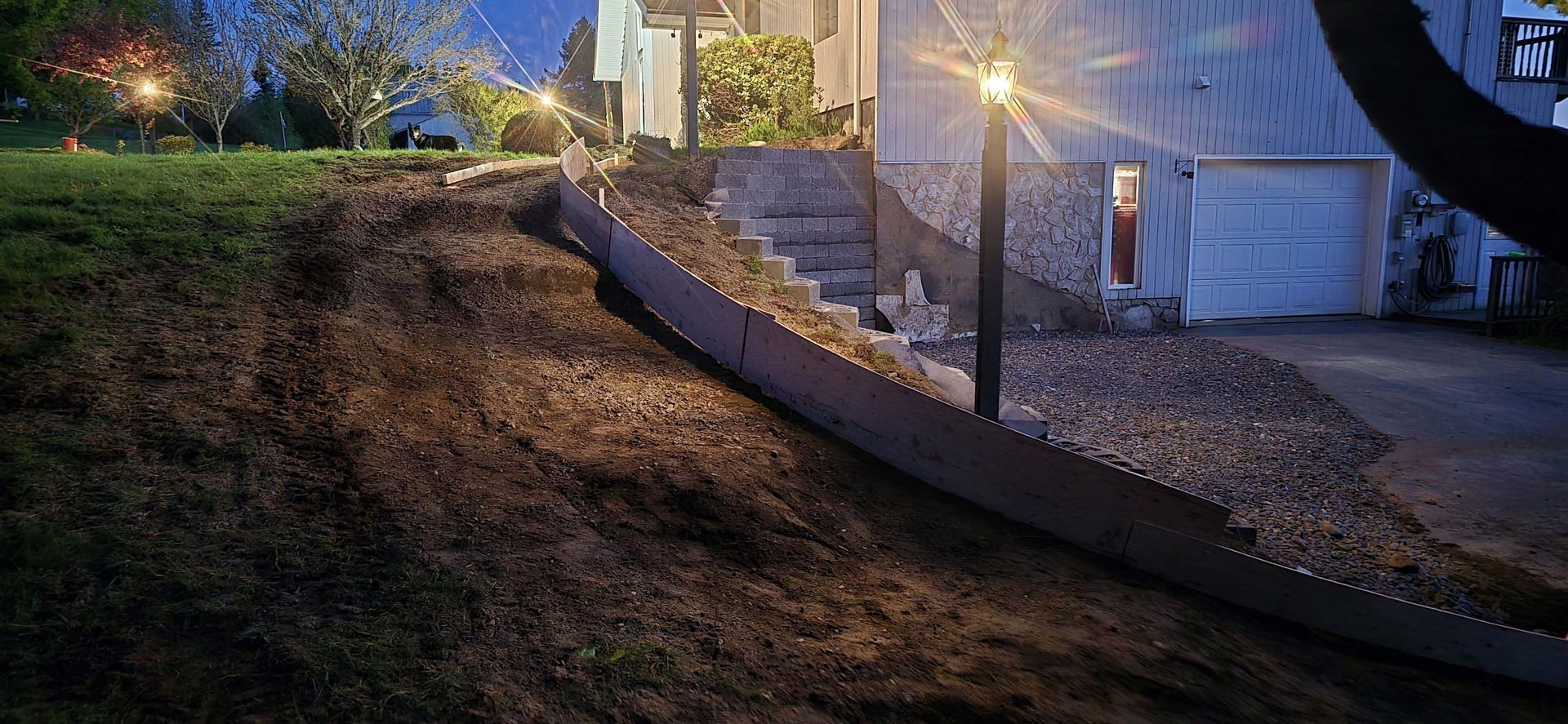 A yard at night features a sloped lawn, a wooden retaining wall, a gravel area, stone steps, and a lit outdoor lamp.