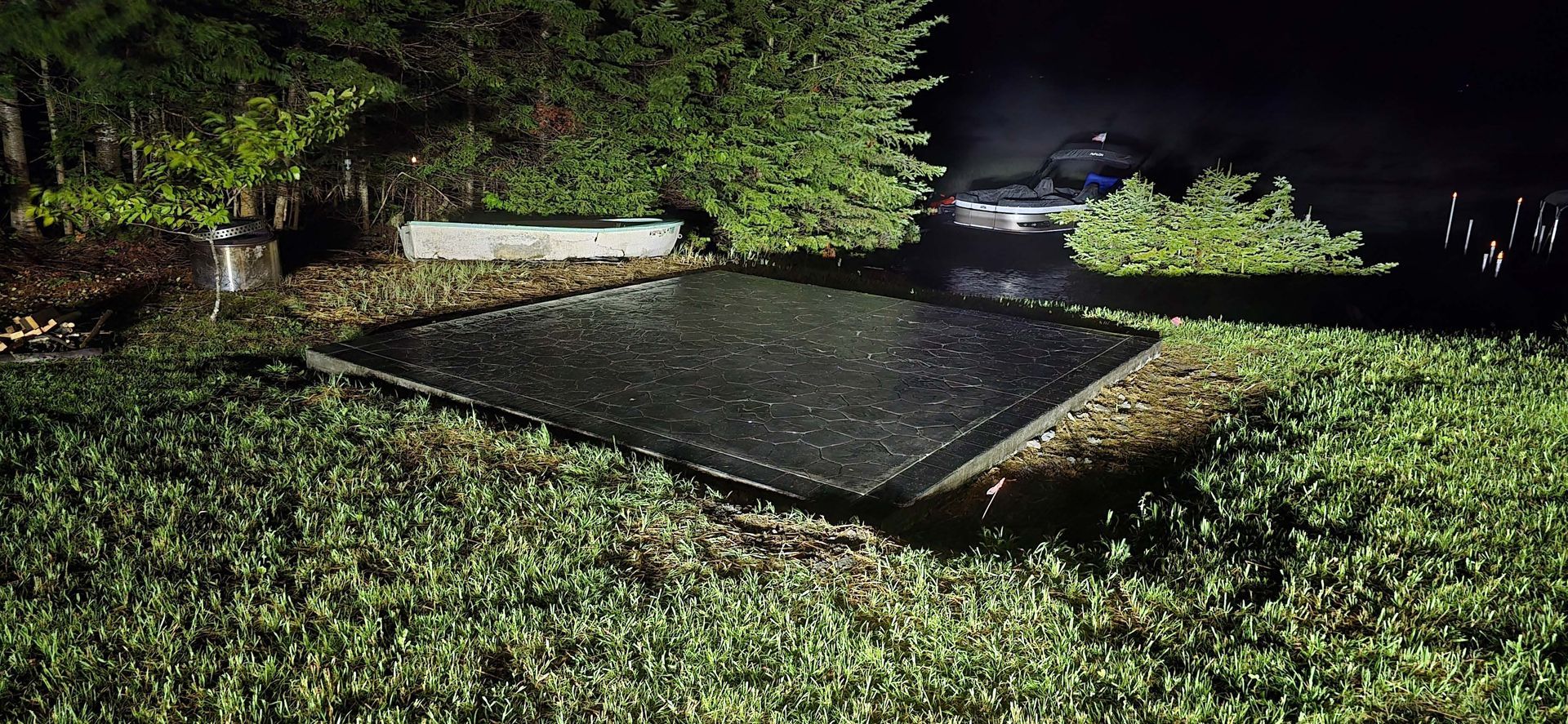 A dark, wet rectangular platform sits on a grassy shore at night near a dark body of water.