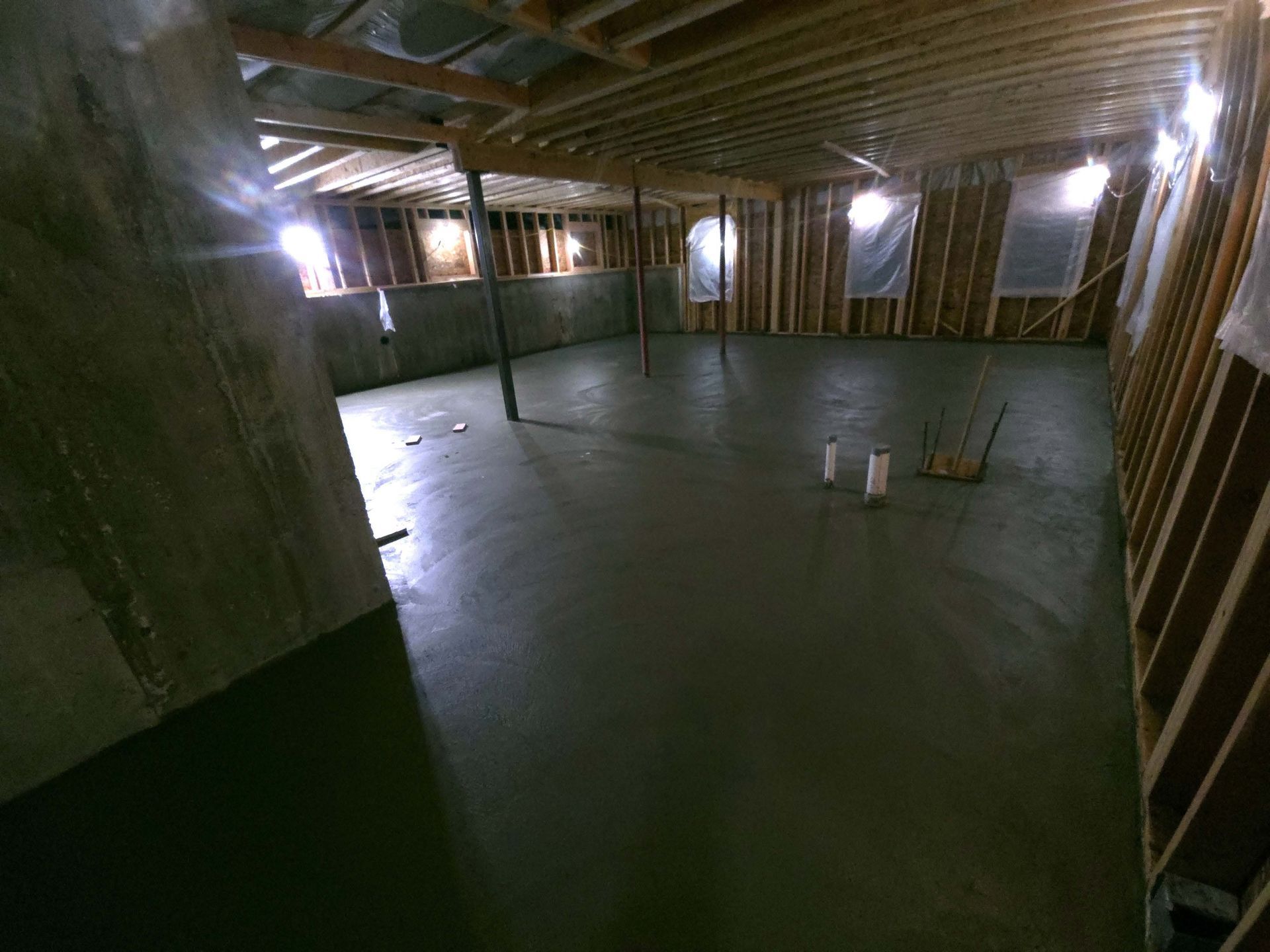 A newly poured concrete basement floor inside a framed wooden structure with support poles and plumbing pipes.