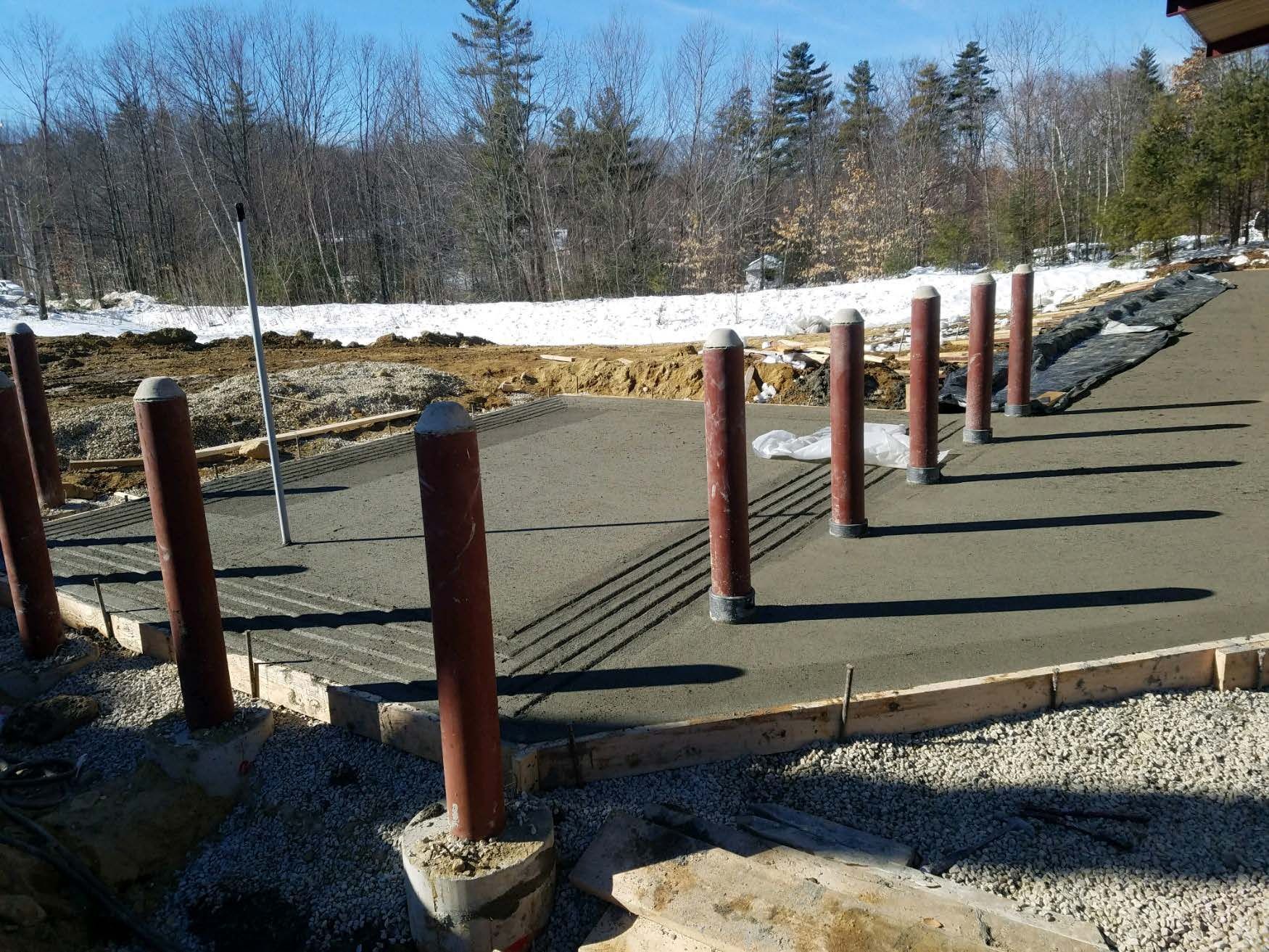 Construction site showing brown metal poles set into fresh concrete footings, surrounded by gravel and wooden forms.