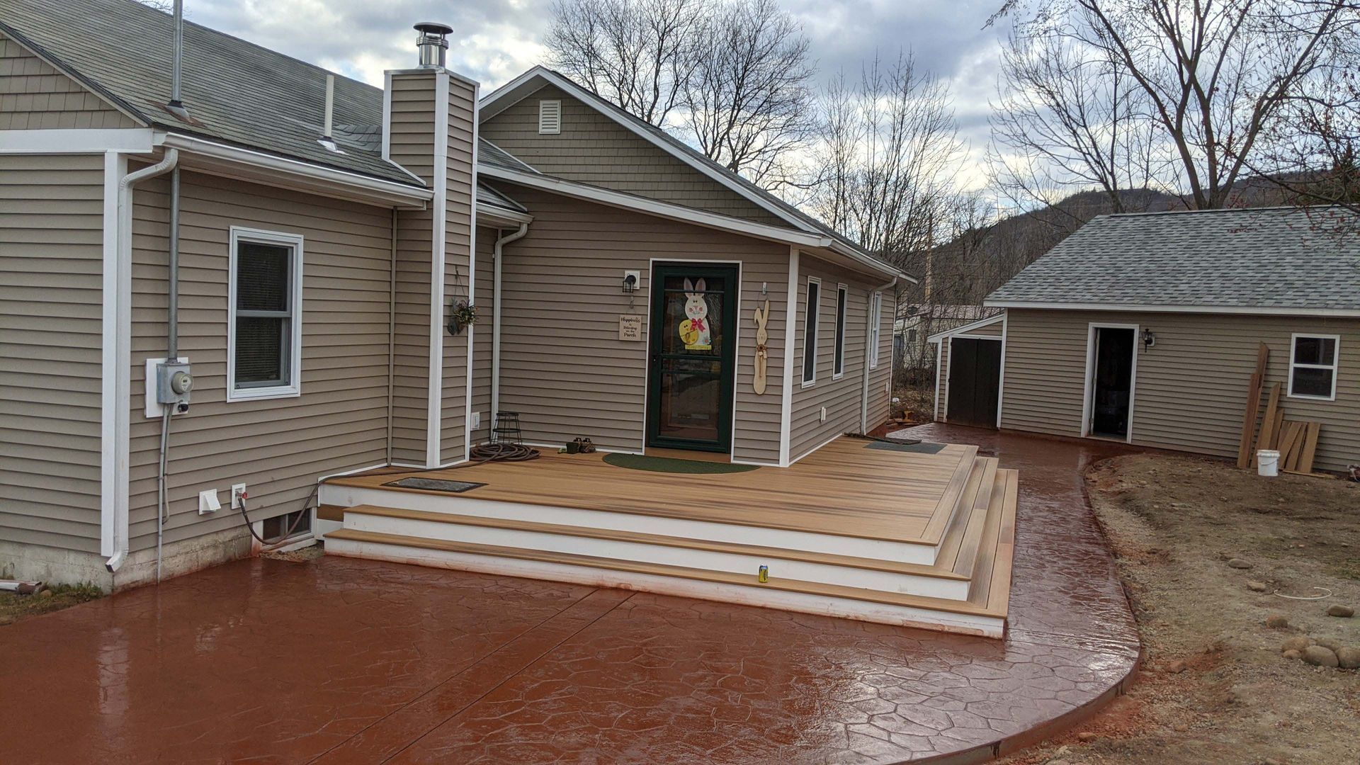 A house with tan siding features a multi-tiered wooden deck and a wet, dark red concrete patio leading to a garage.