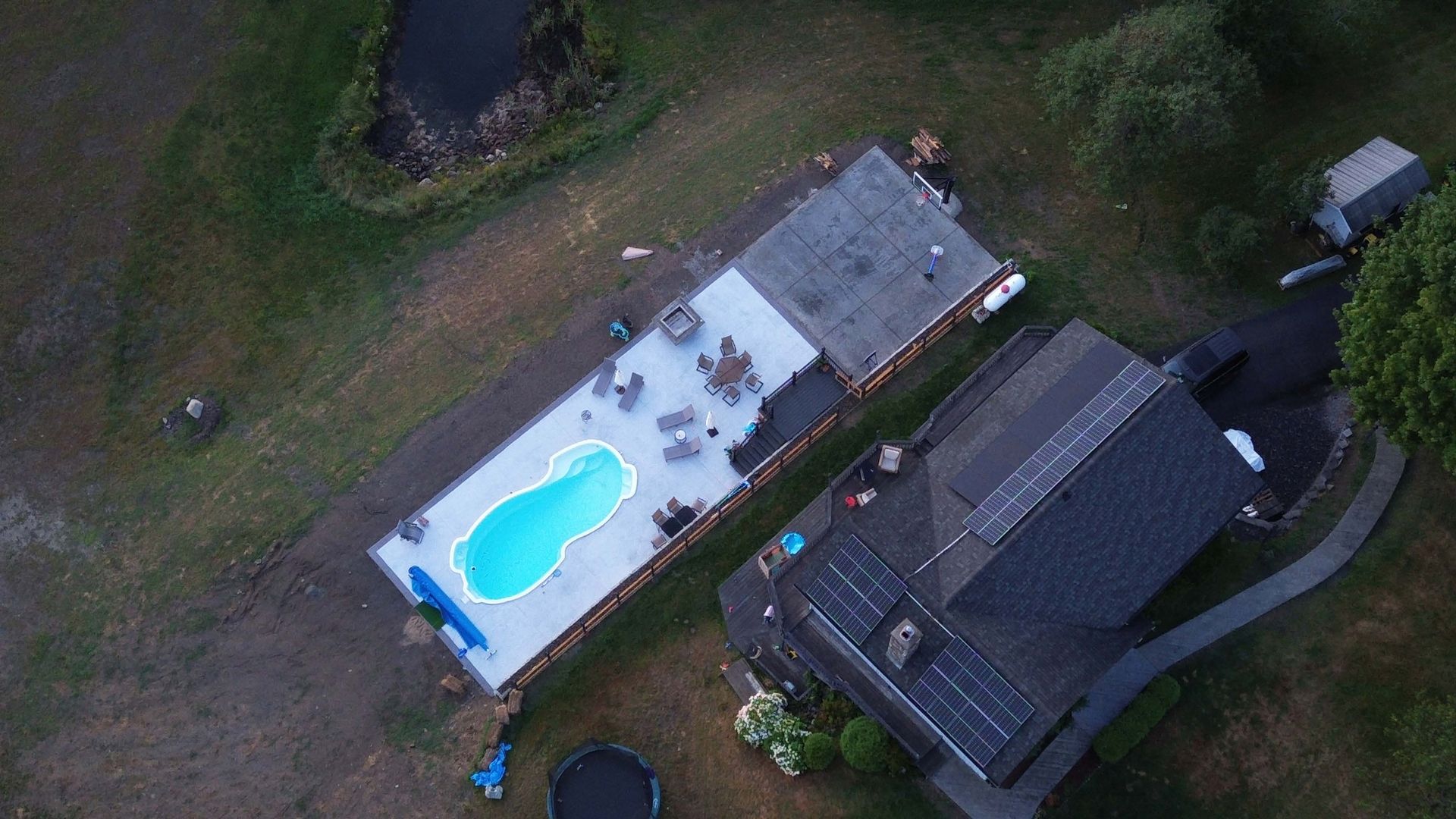 Aerial view of a backyard with a house, a detached garage with a flat roof, and an in-ground pool on a paved patio.