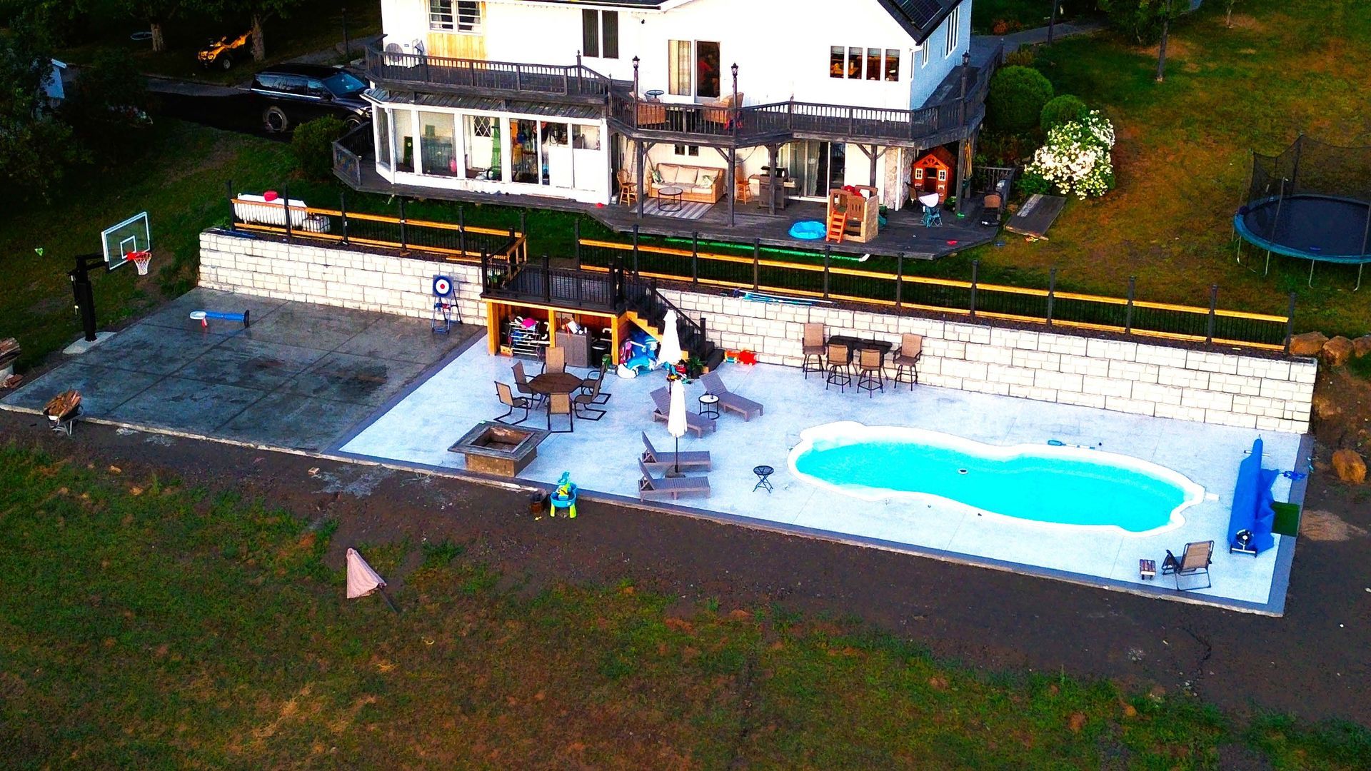 Aerial view of a backyard with a house, a paved patio, a swimming pool, a basketball hoop, and a small play structure.