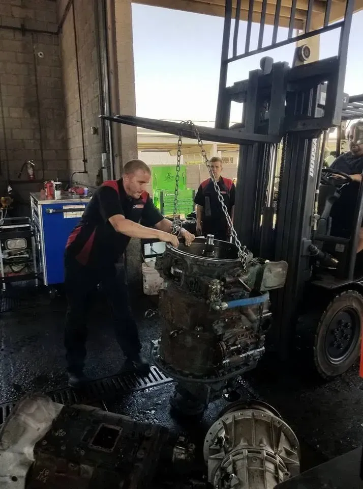 Mechanic using forklift to lift engine in garage, two others nearby.