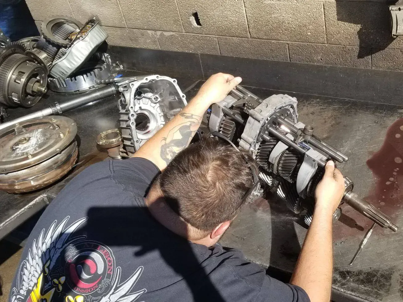 Mechanic disassembling a car transmission on a workbench; parts and tools are scattered around.