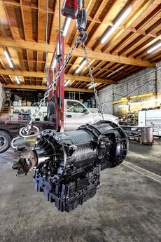 A large, black transmission is suspended from a crane in a workshop.