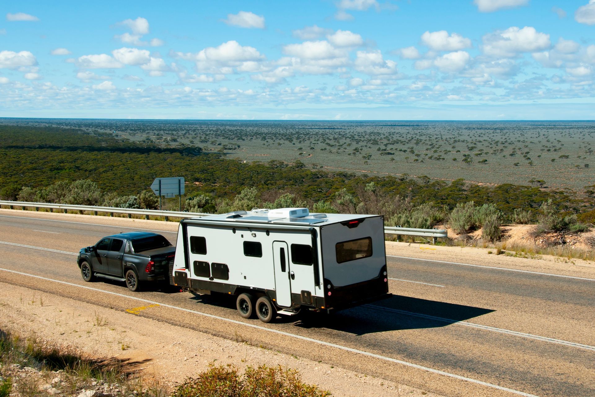 Black truck towing a white and black RV on a highway through a semi-arid landscape under a blue sky.