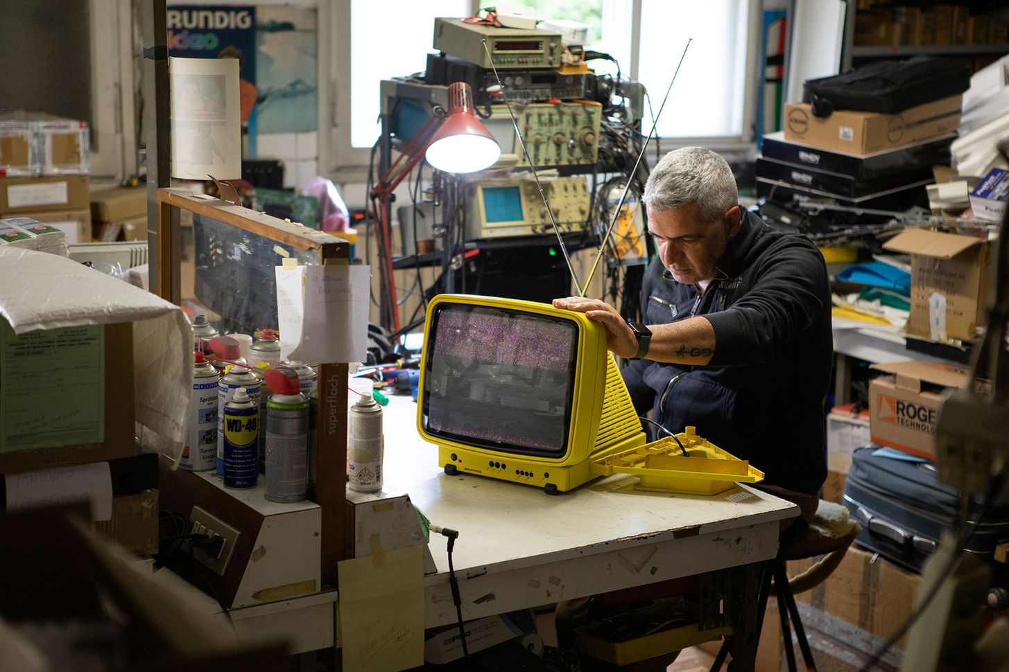 Person working on a yellow machine in a cluttered workshop, surrounded by tools and shelves.