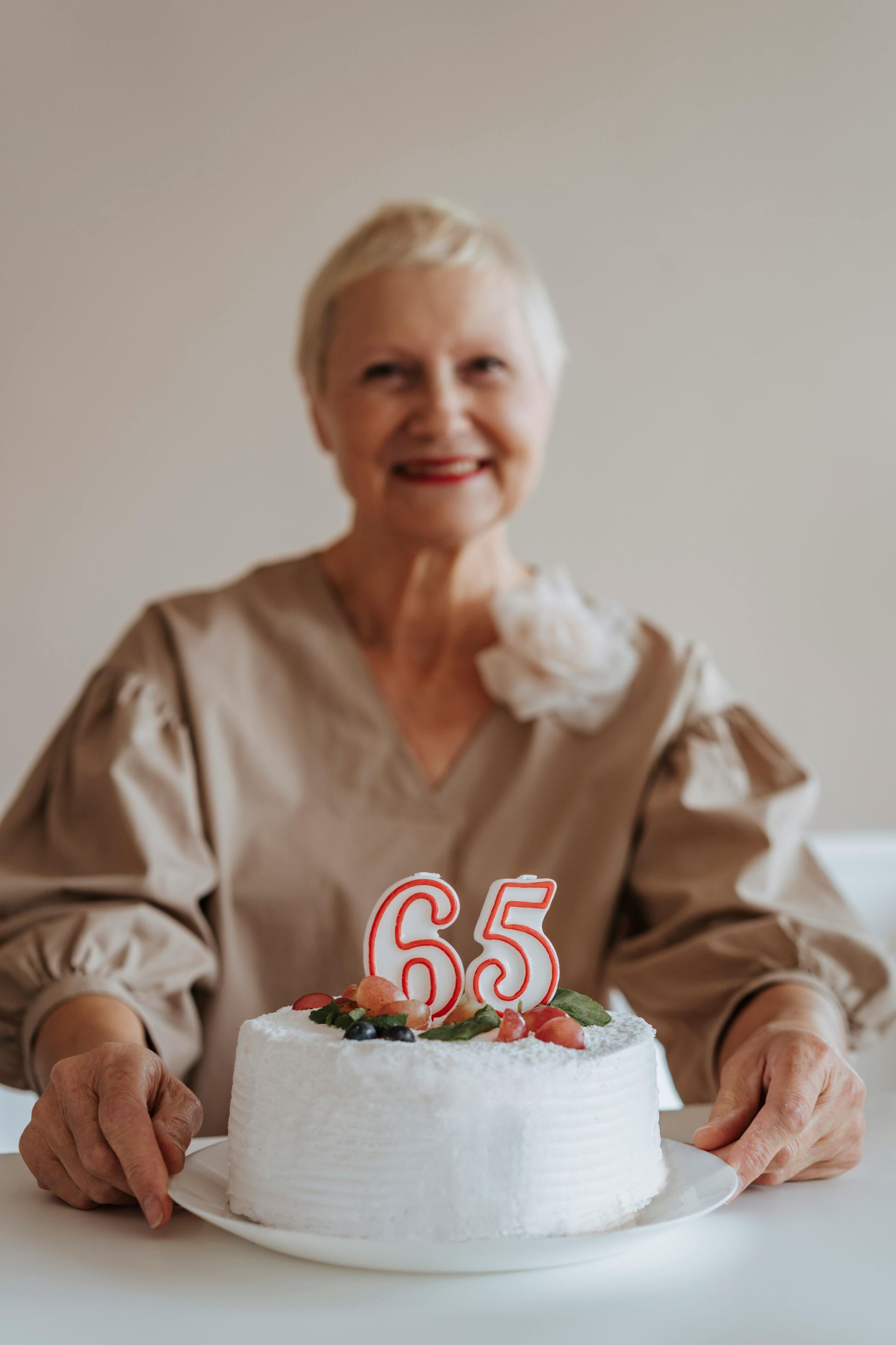 Woman smiling, holding a white birthday cake with 65 candles on a table.