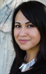 Woman with dark hair smiles, wearing a black shirt and silver necklace. Leans against a light-colored, weathered background.
