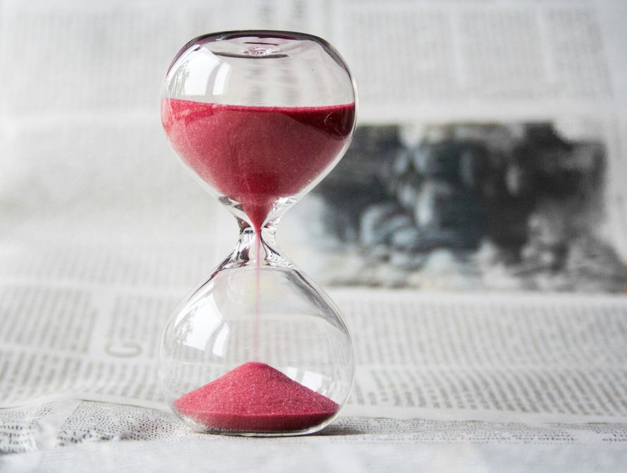 Hourglass with red sand, nearly run out, set against a blurred newspaper background.