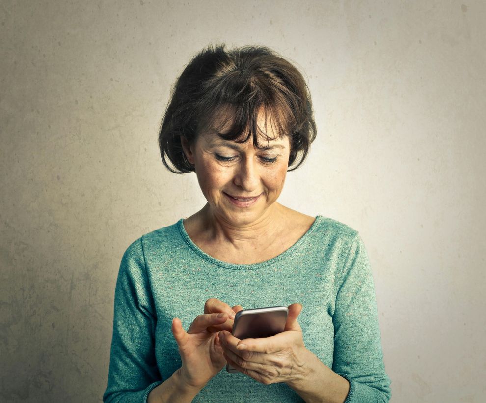 Smiling older woman using a smartphone; teal shirt, neutral background.