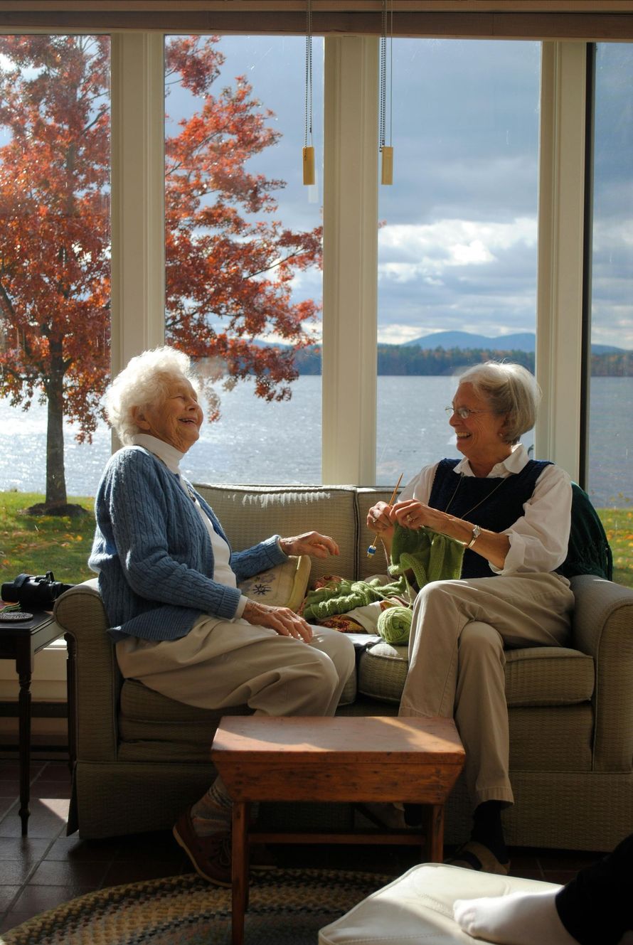 Two older women laughing on a sofa by a large window overlooking a lake with fall foliage.