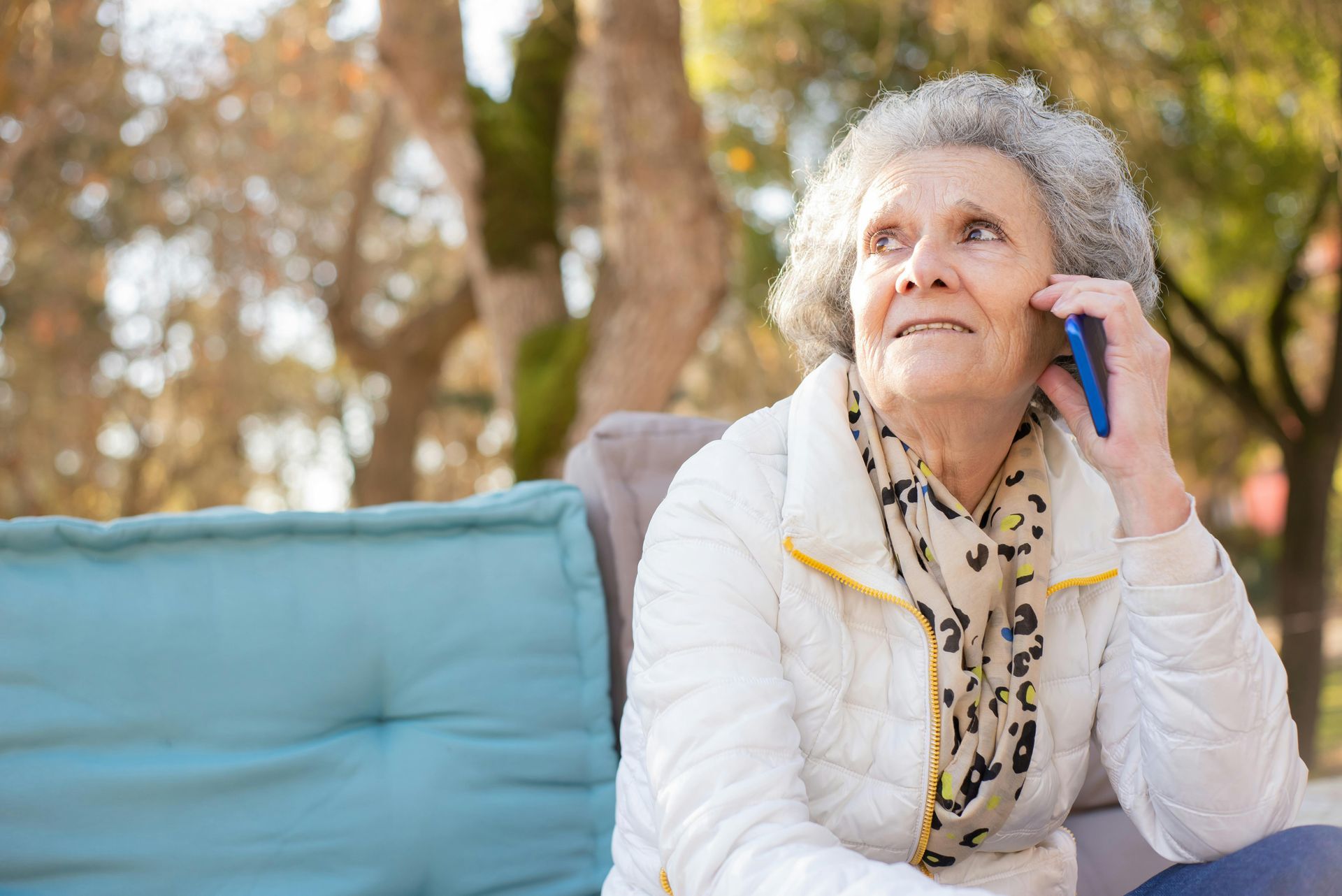 Elderly woman with gray hair smiles while talking on her phone outdoors, seated on a cushioned bench.