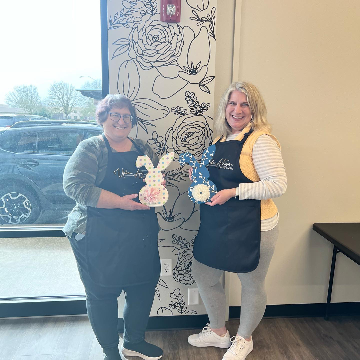 Two women are standing next to each other in front of a wall with flowers on it.