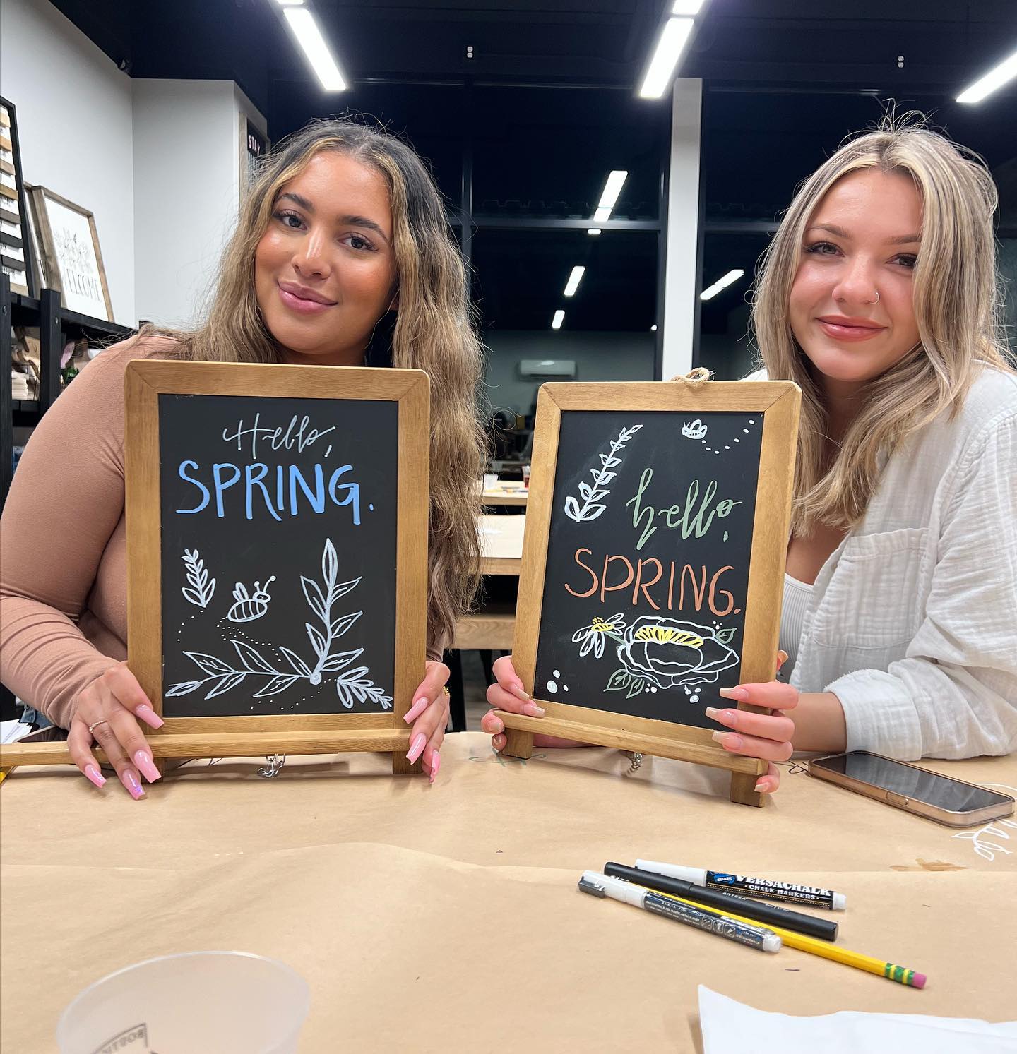 Two women are sitting at a table holding chalkboards that say hello spring.