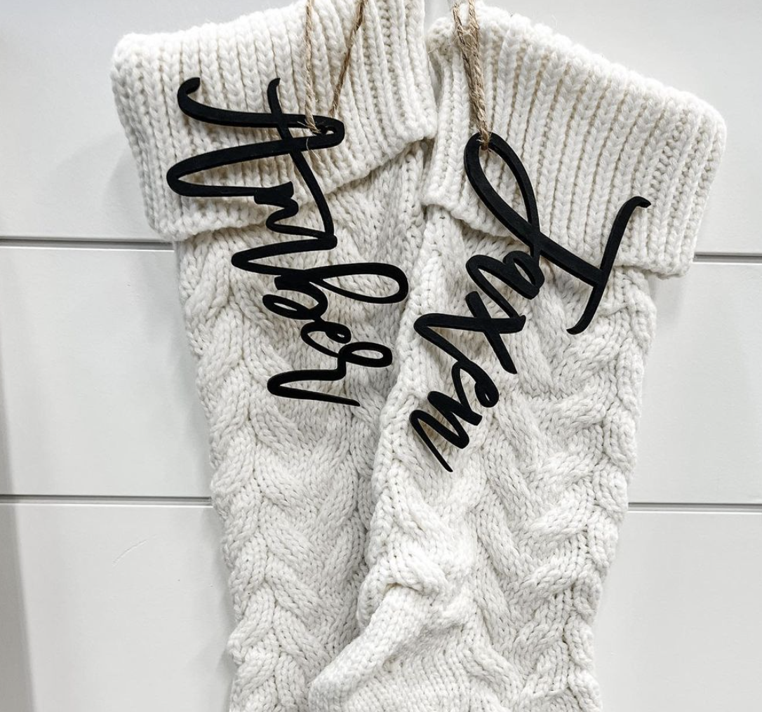 A pair of white knitted socks with black writing on them hanging on a white wall.
