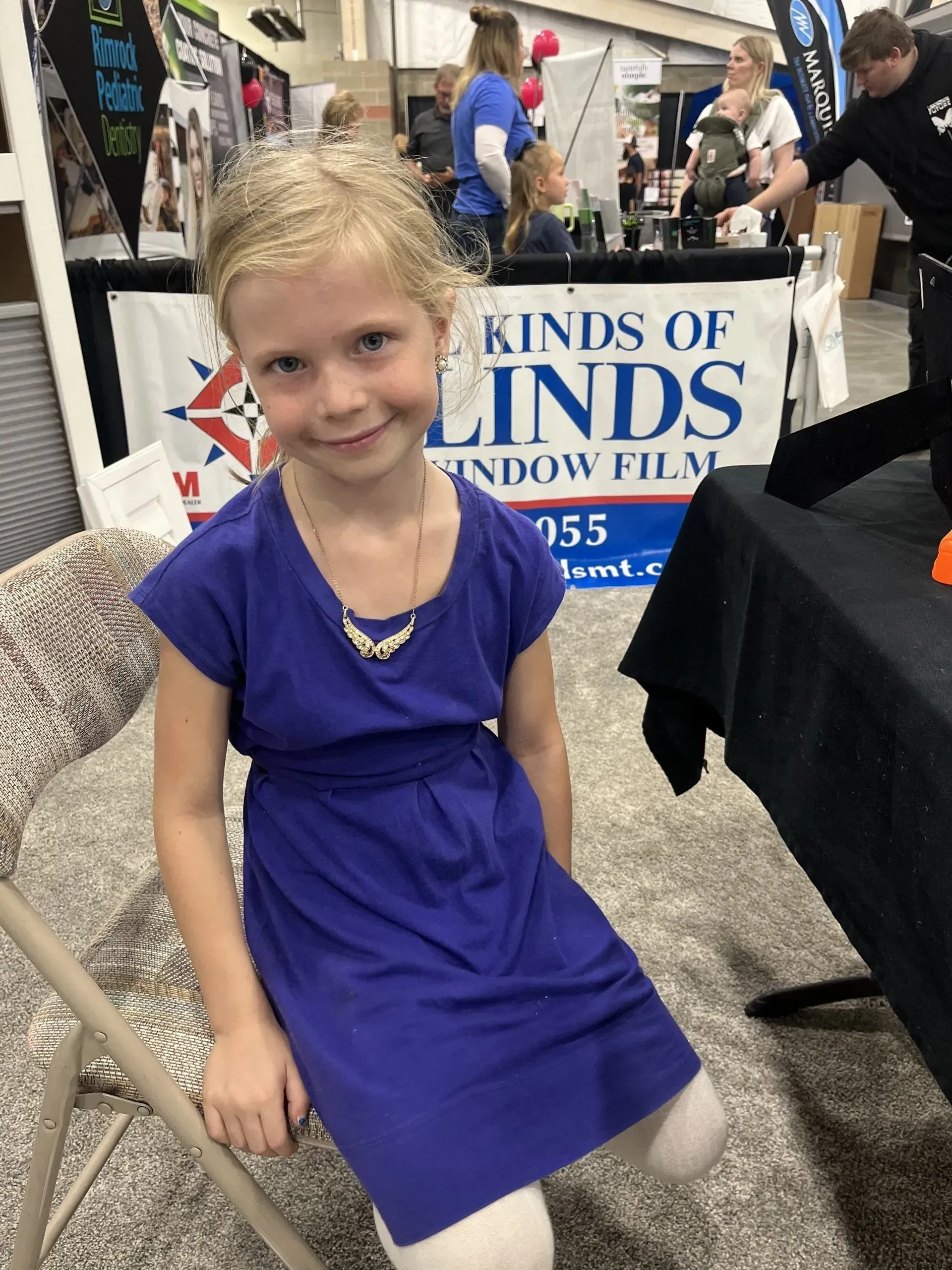 A little girl in a purple dress is sitting in front of a kinds of blinds sign.