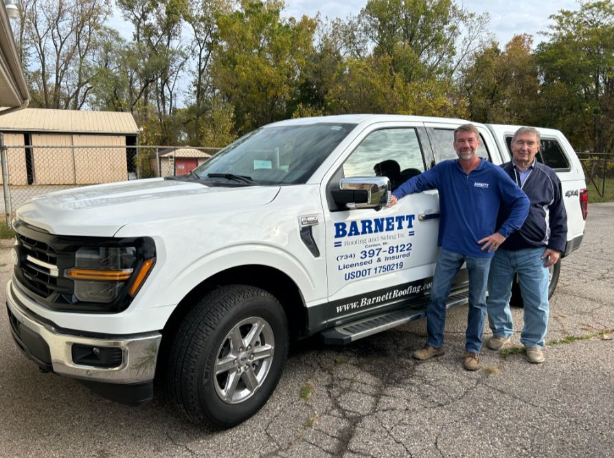 Two men pose next to a white Barnett truck. One man wears blue, the other a jacket. Outdoors, trees in background.