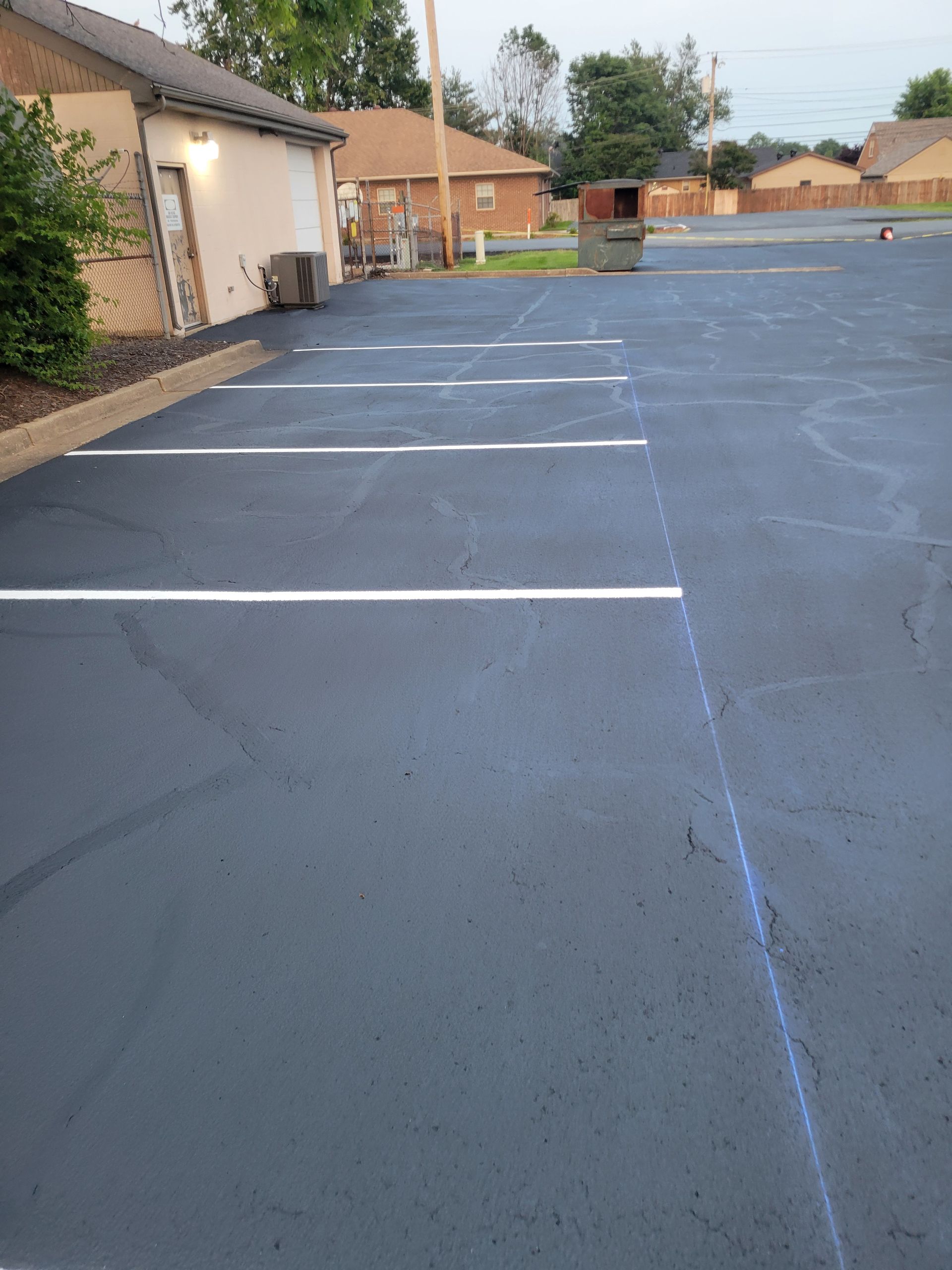 A parking lot with a house in the background and a trash can in the middle.