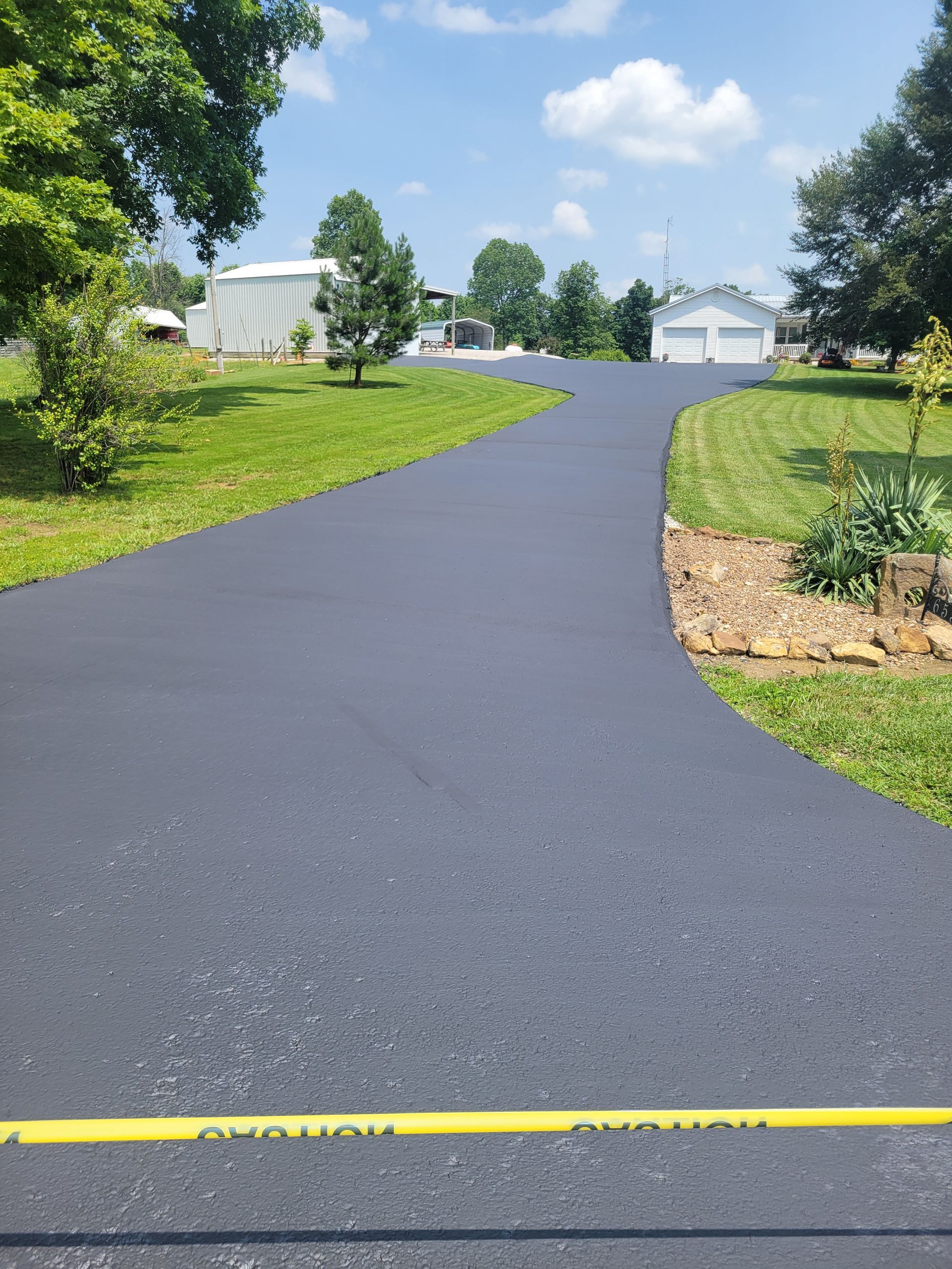 Newly paved asphalt driveway curving towards a garage on a sunny day, with green lawn and trees flanking.