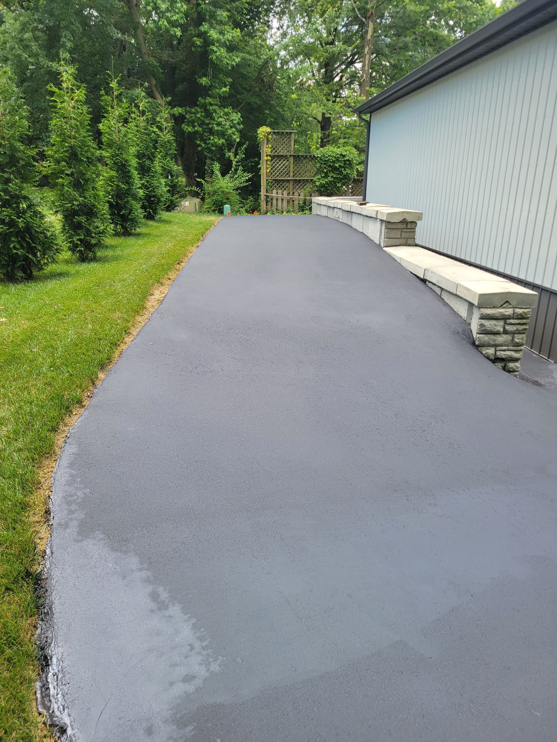 Newly paved black asphalt driveway curving toward a wooden structure, bordered by green grass and evergreen trees.