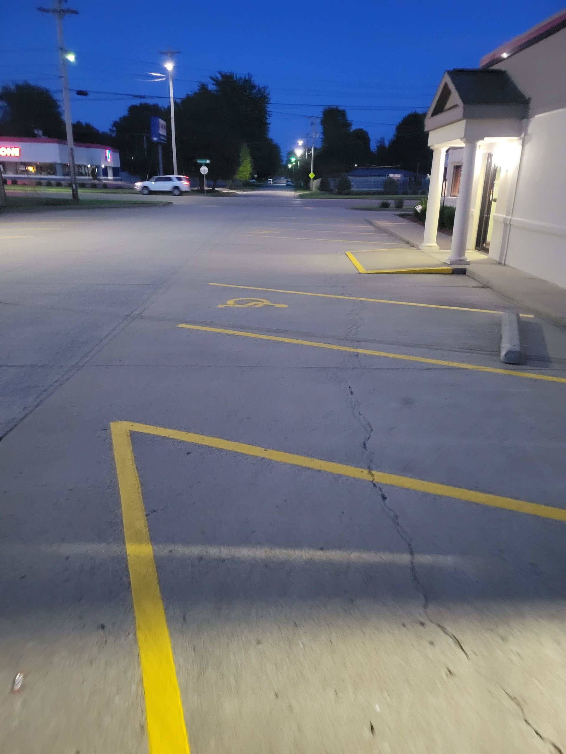 Nighttime view of a nearly empty parking lot next to a building with bright lights over an entrance. Yellow parking space lines are visible.