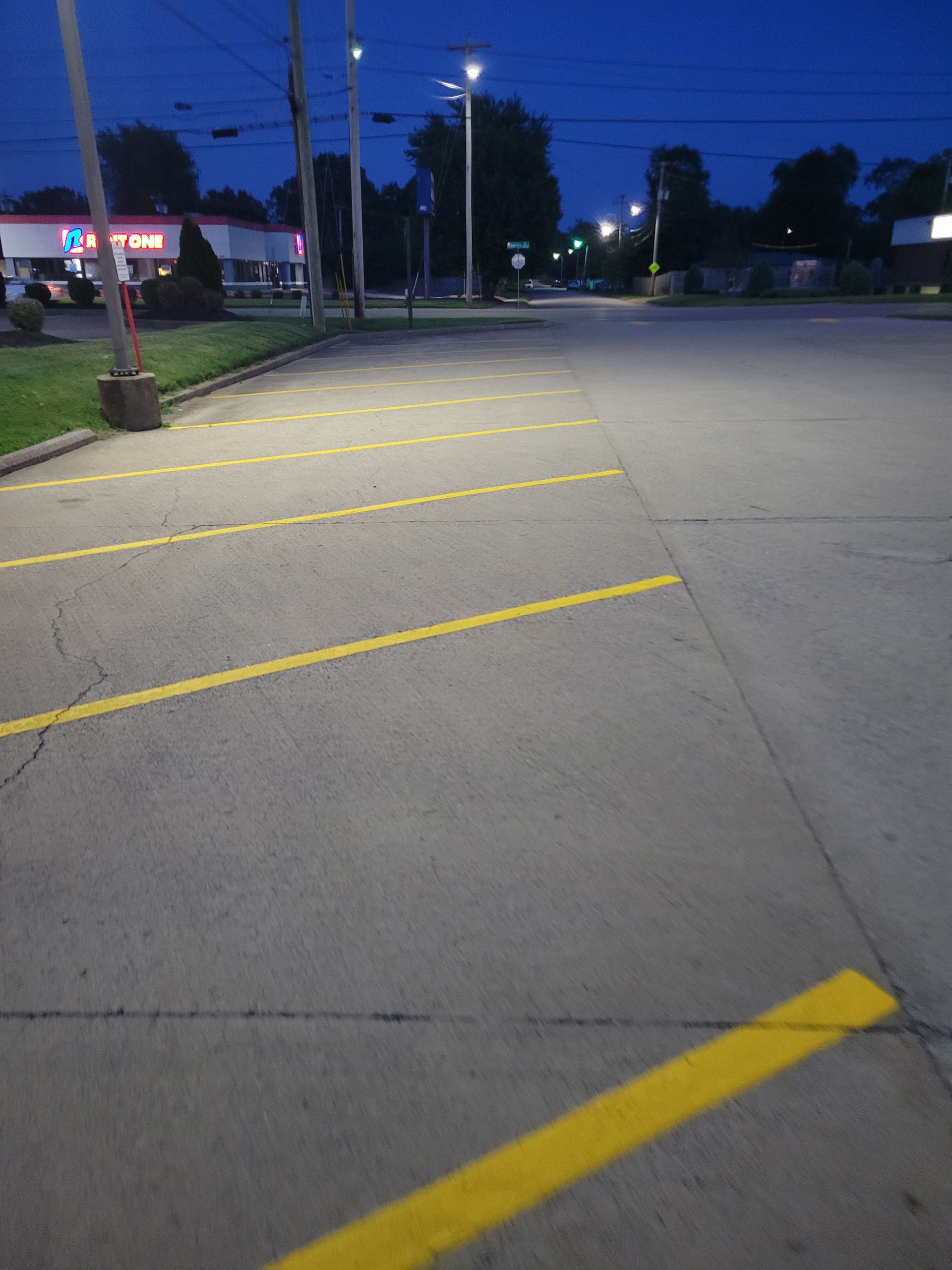 Empty parking lot at dusk. Yellow parking lines on gray pavement. Streetlights illuminate the scene.