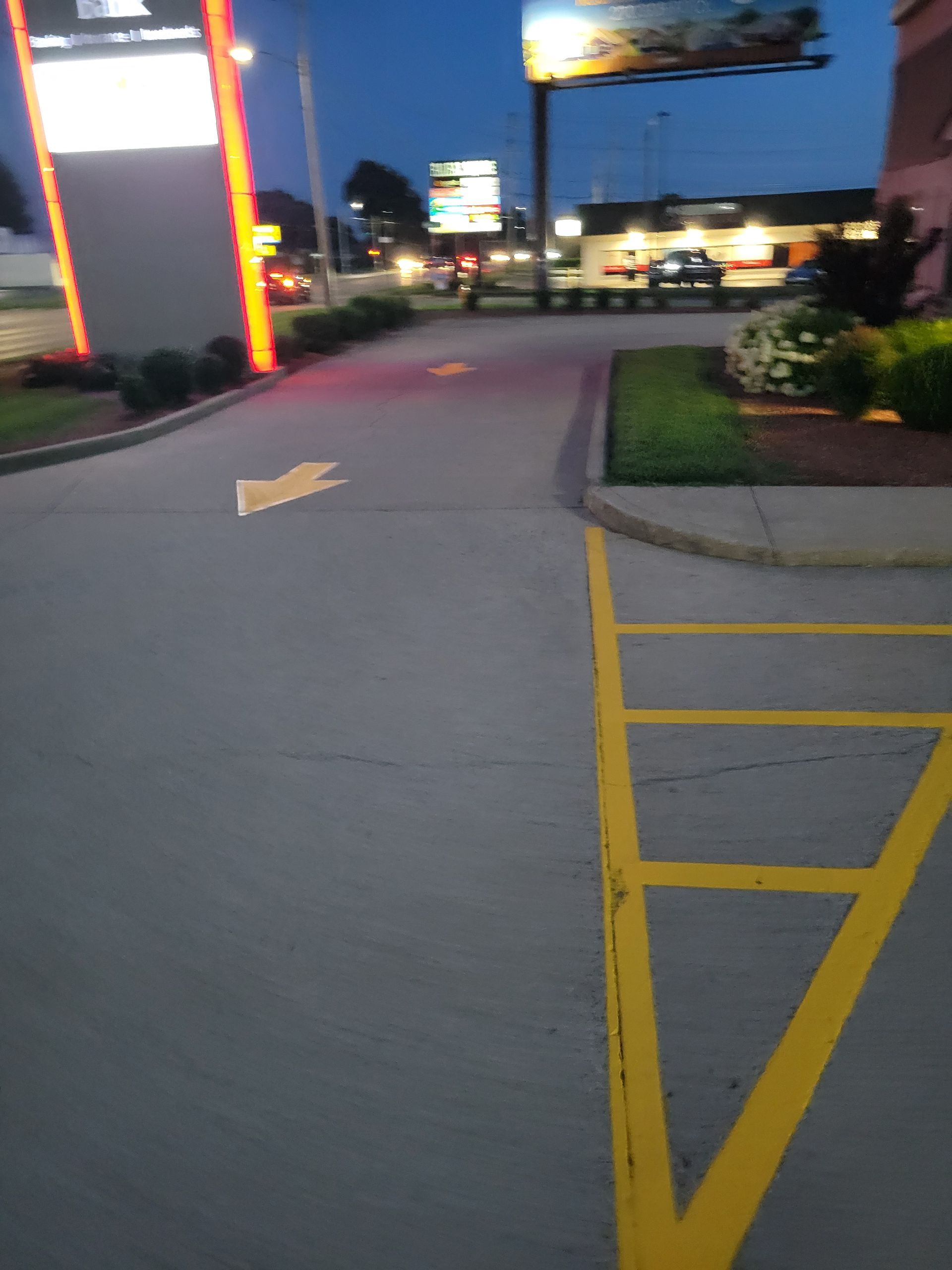 Drive-thru lane at dusk, with yellow lines and an arrow guiding cars. A bright sign and building are visible.