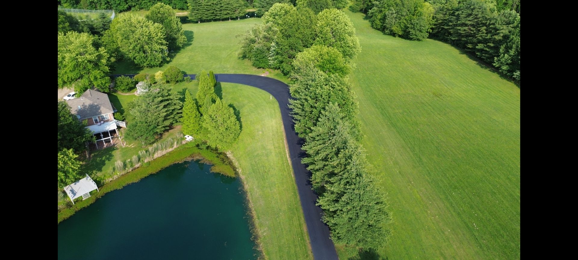 Aerial view of a house on a lake. A paved road winds through a grassy field with trees.