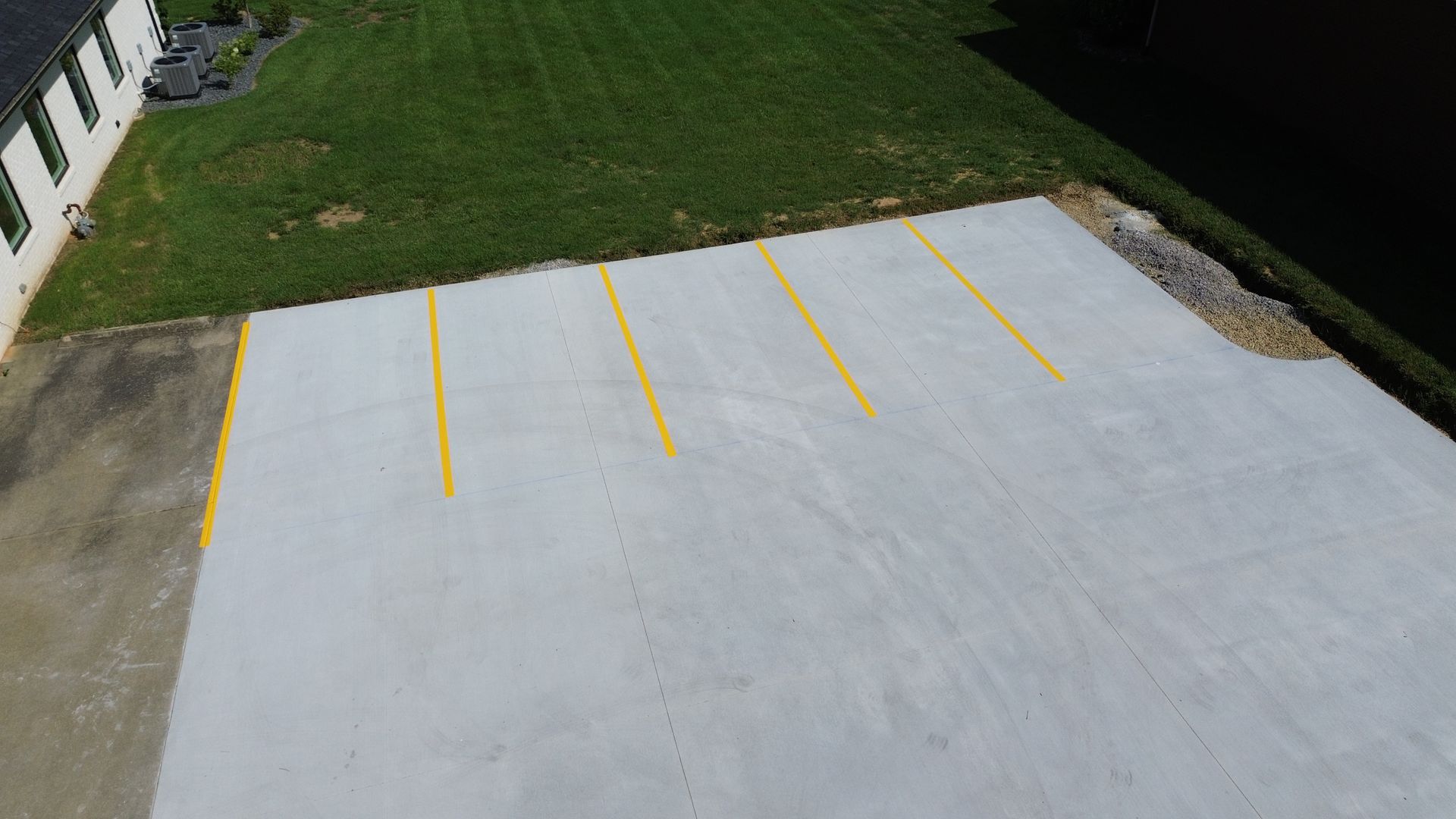 Overhead view of a newly paved parking lot with yellow painted lines, adjacent to a building and grass lawn.