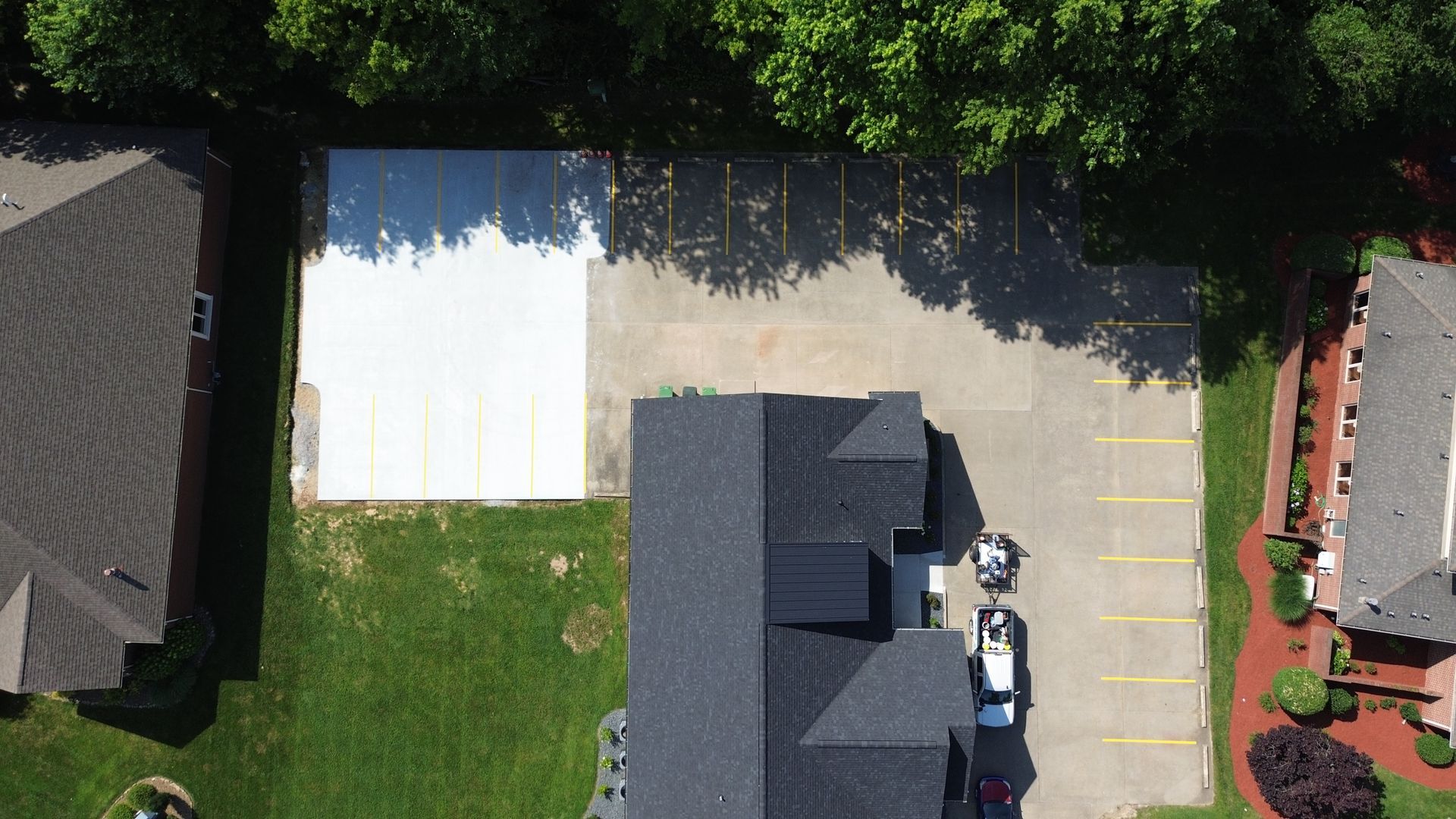 Overhead view of a building with a dark roof and cars parked in front. A white, square structure sits next to it, with trees to the right.