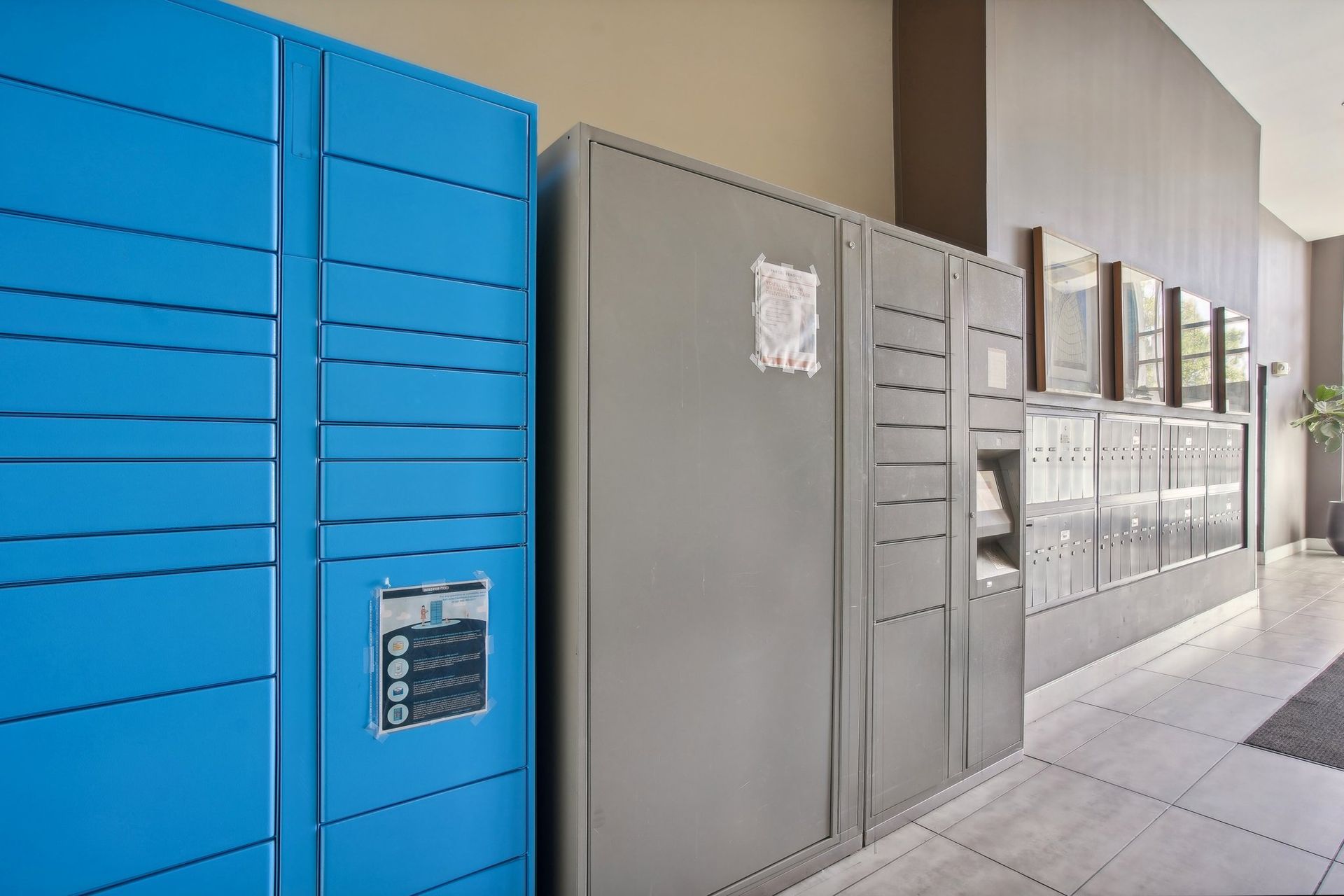 Blue and gray metal package lockers and mailboxes in a well-lit hallway.
