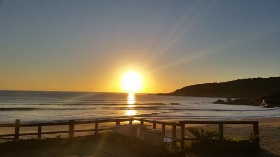 Um sol brilhante se põe sobre uma praia oceânica tranquila, emoldurada por uma cerca de madeira em primeiro plano.