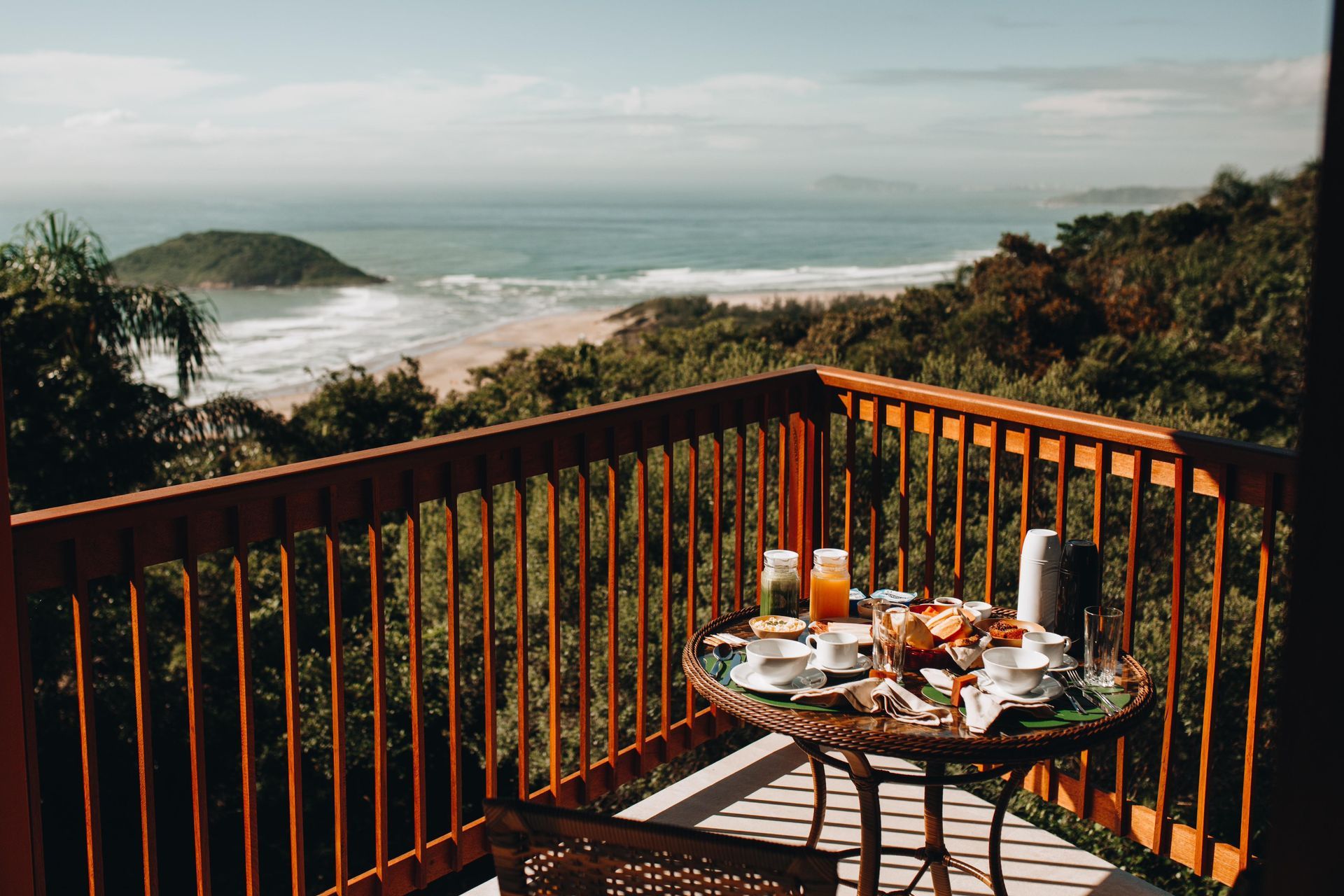 Um café da manhã servido em uma mesa na varanda com vista para uma praia de areia, o oceano e a exuberante vegetação costeira sob um céu azul límpido.