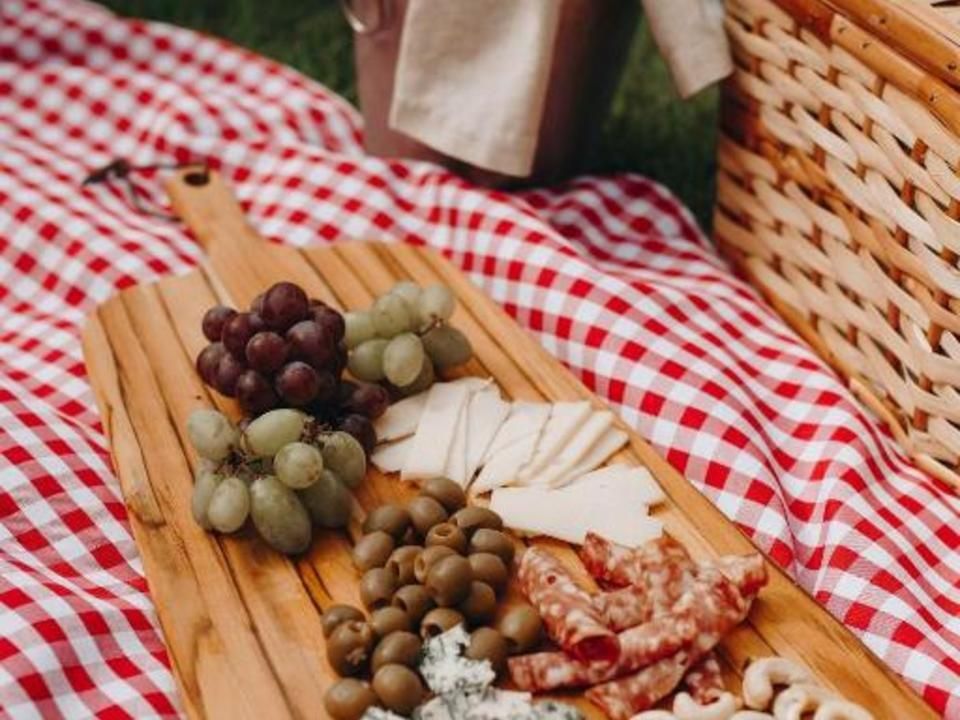 Uma tábua de charcutaria com uvas, azeitonas, queijo e salame repousa sobre uma toalha de piquenique xadrez vermelha e branca.