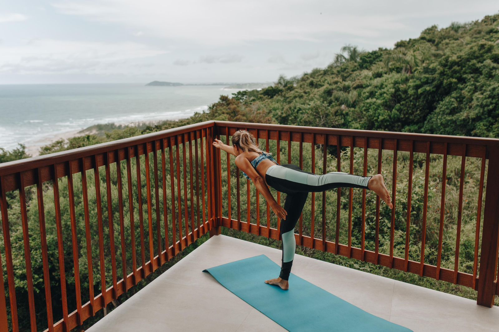 Uma pessoa executa a postura do guerreiro de ioga em um tapete azul em uma varanda com vista para o oceano e as colinas.
