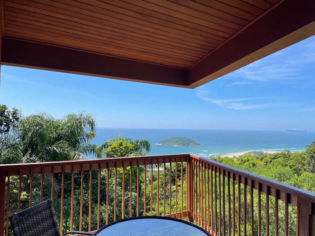 Uma varanda de madeira com vista para uma exuberante floresta verde, uma pequena ilha e o oceano azul sob um céu límpido.