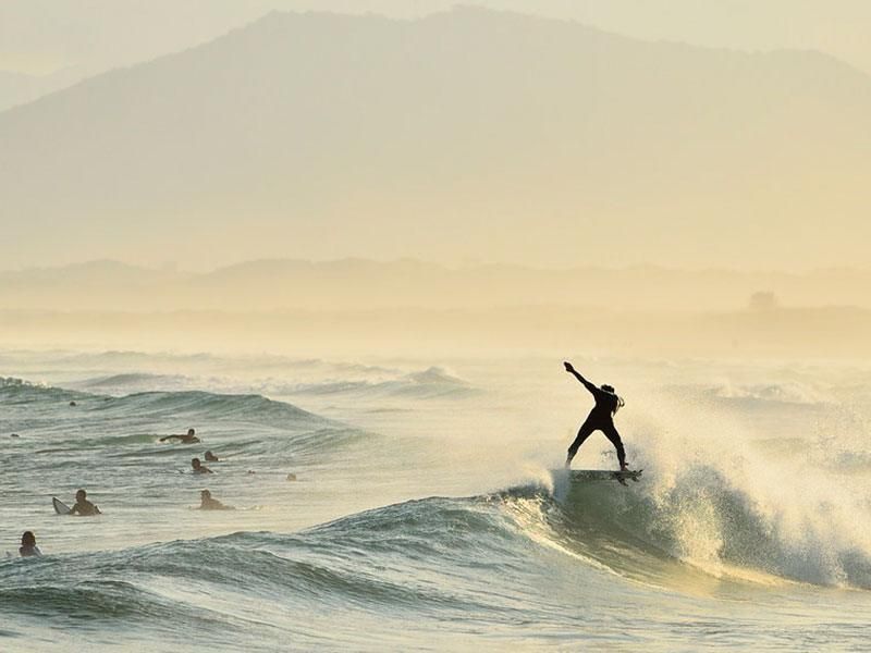 Um surfista em pose dinâmica desliza sobre uma onda durante um pôr do sol enevoado, com outros nadadores visíveis na água próxima.