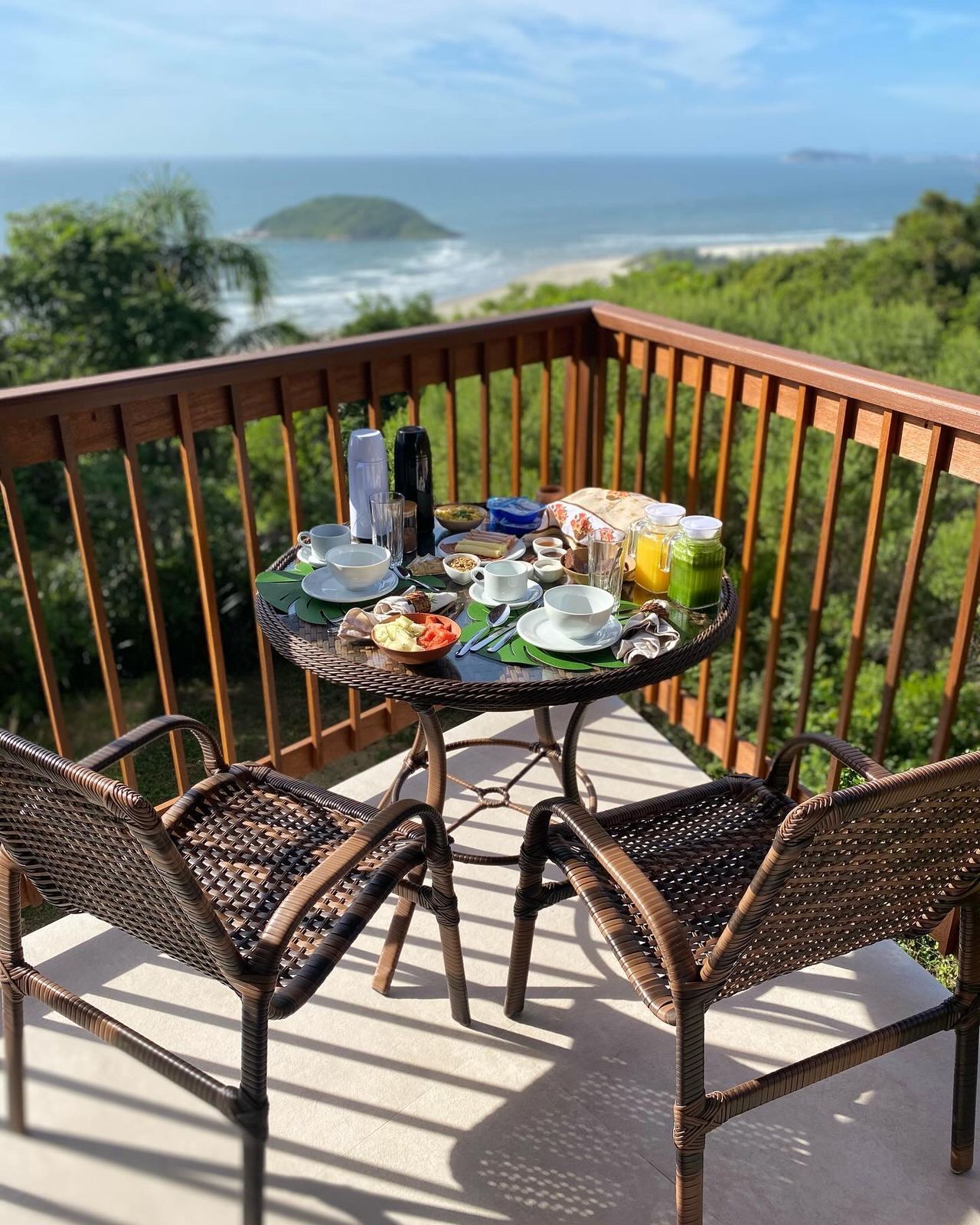 Uma mesa posta para dois com itens de café da manhã em uma varanda com vista para uma praia oceânica deslumbrante e vegetação exuberante.