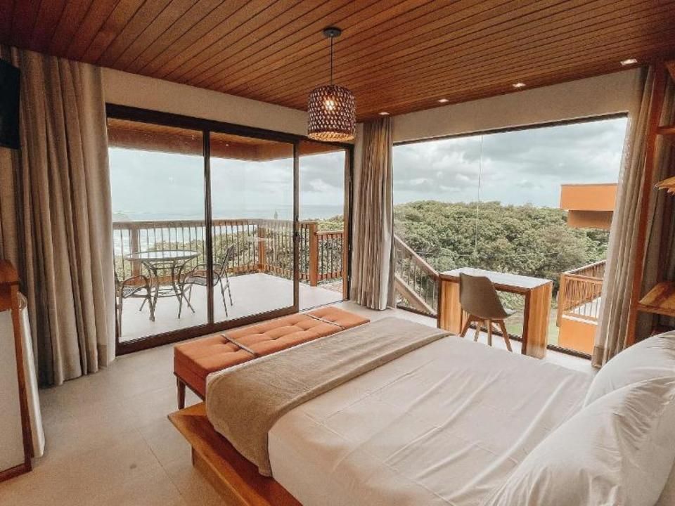 Um quarto com teto de madeira, uma cama de casal, uma escrivaninha e portas de vidro do chão ao teto que se abrem para uma varanda com vista.