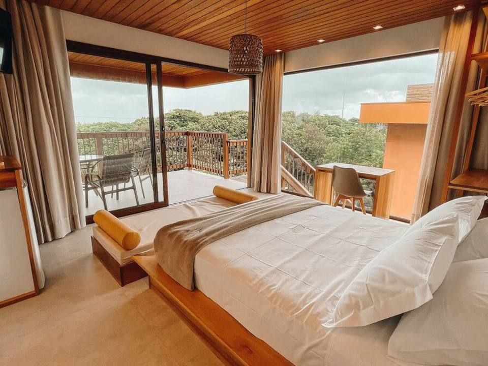 Quarto moderno com cama plataforma, teto de madeira e grandes portas de vidro que se abrem para uma varanda arborizada com vista panorâmica.