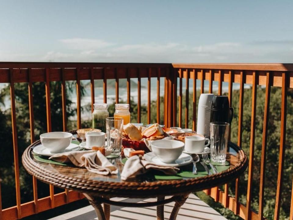 Um café da manhã posta com tigelas brancas, canecas e frutas sobre uma mesa redonda em uma varanda de madeira com vista para o oceano.