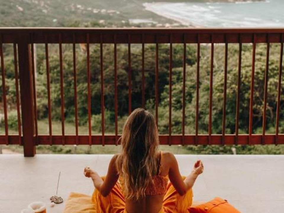 Uma pessoa está sentada em posição meditativa sobre uma almofada laranja em uma varanda, com vista para uma paisagem exuberante e para o oceano.