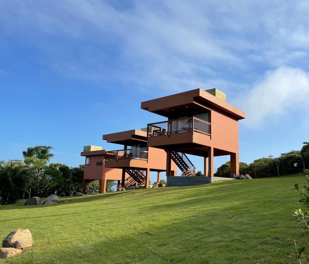 Duas cabanas modernas, elevadas e de cor ferrugem, com varandas, estão situadas em uma colina gramada sob um céu azul claro.
