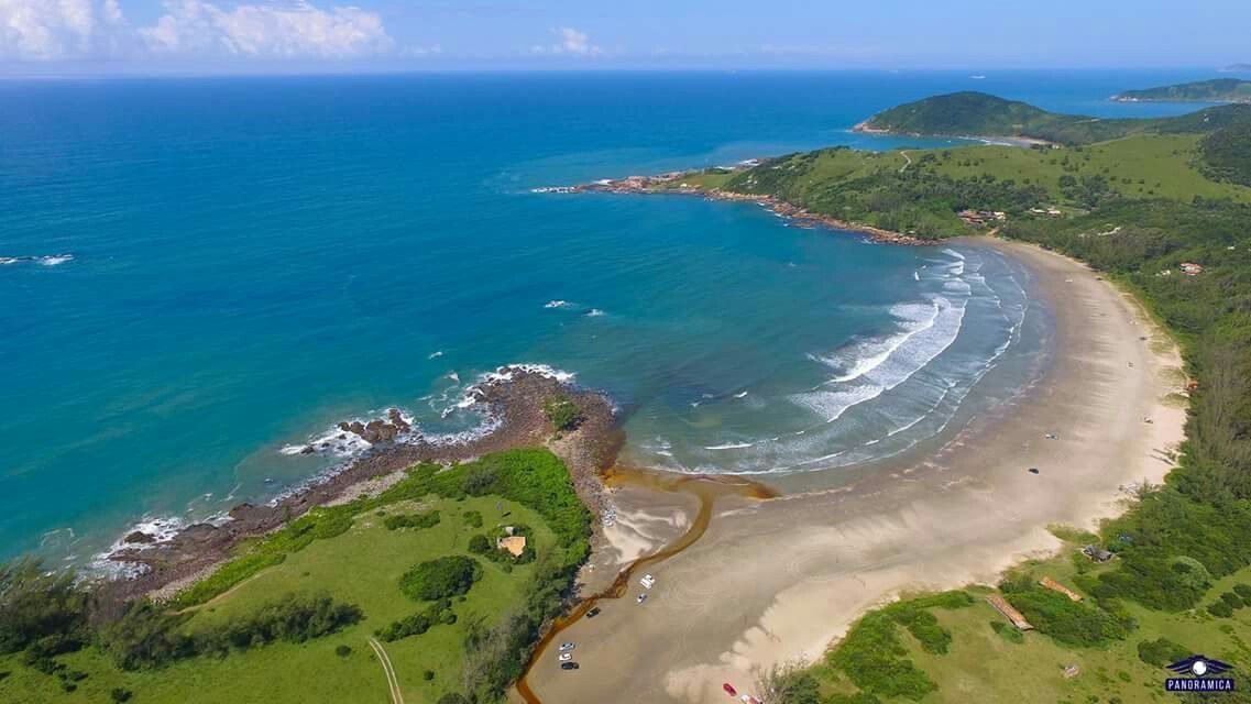 Vista aérea de uma praia de areia curva que encontra colinas verdejantes e onduladas com as águas turquesas do oceano.