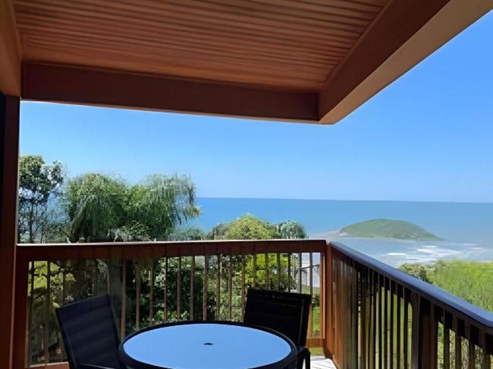 Uma varanda com uma pequena mesa e duas cadeiras com vista para uma paisagem oceânica deslumbrante e uma ilha gramada ao longe.